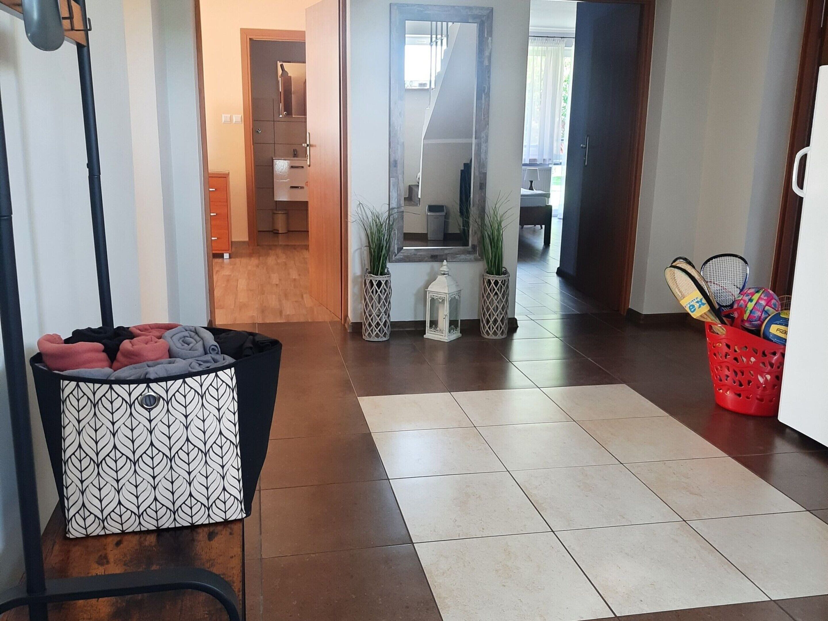 A hallway with a patterned trash bin, a red basket with sports equipment, and a tiled floor leading to a room with a mirror and a plant.