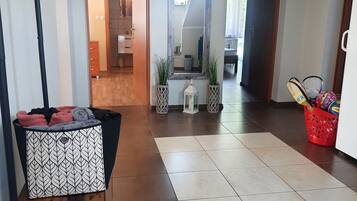 A hallway with a patterned trash bin, a red basket with sports equipment, and a tiled floor leading to a room with a mirror and a plant.