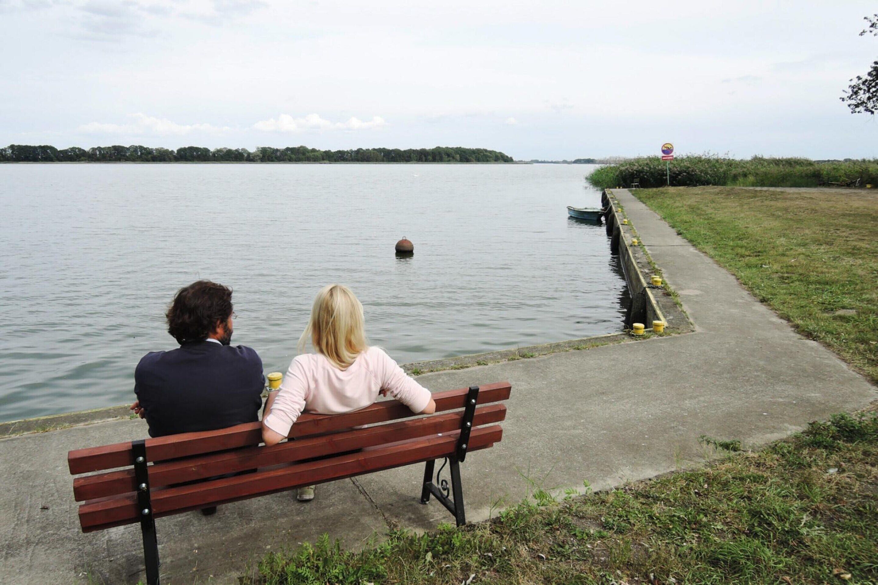 Twee personen zitten op een bankje naast kalm water, met een grasveld en een betonpad dat naar een klein bootje leidt.