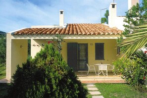 A small, single-story building with a tiled roof, a door with a window, and a small patio area with a table and chairs. - Apartment on Costa Rei Near Sandy Beaches (Muravera)