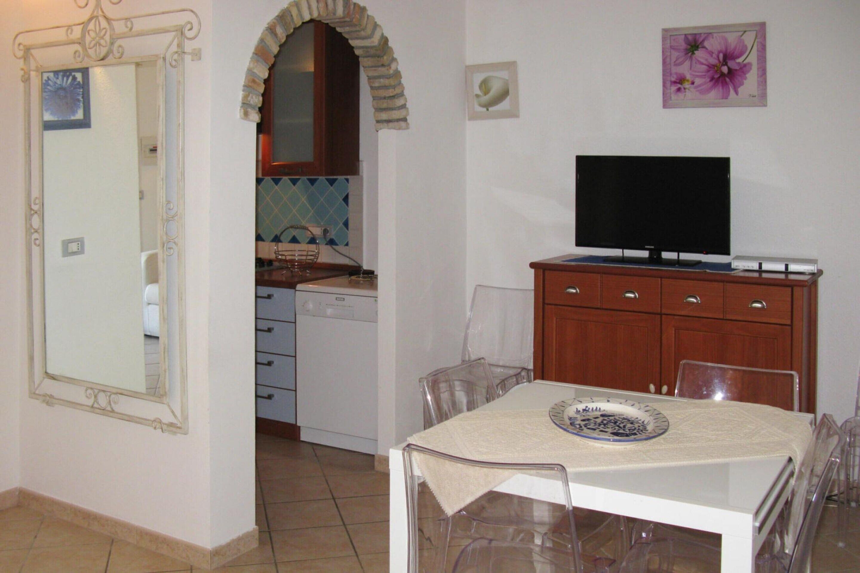 Una cocina compacta con lavavajillas, una mesa de comedor con sillas, un televisor sobre un mueble de madera y un espejo decorativo.