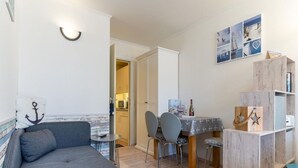 A cozy living room with a grey sofa, a small dining table with chairs, and a wooden bookshelf. - Ferienwohnung, St. Peter - Ording (St. Peter-Ording)
