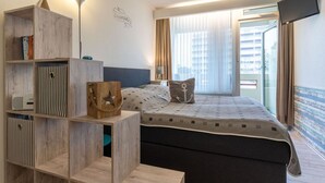 A hotel room with a bed, a wooden shelf unit, a flat-screen TV, and a window with curtains. - Ferienwohnung, St. Peter - Ording (St. Peter-Ording)