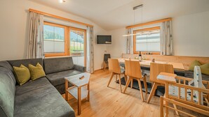Ett modernt vardagsrum med en grå soffa, ett matbord i trä och stolar. Det finns stora fönster med gardiner, en platt-TV och en taklampa.