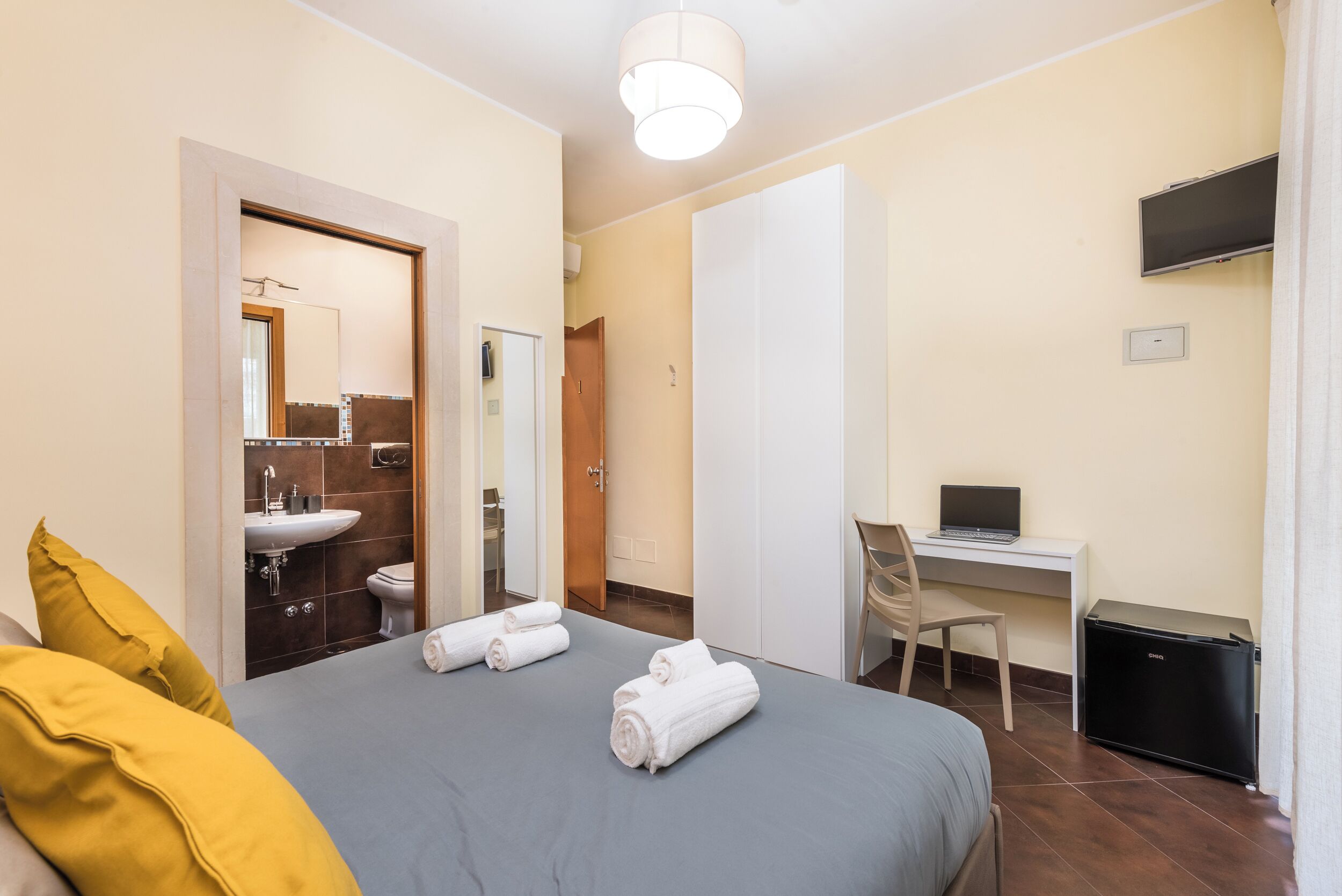 Une chambre d’hôtel comprenant un lit, un bureau avec un ordinateur portable, un petit réfrigérateur, une chaise, une salle de bain avec un lavabo et des toilettes, et un miroir.