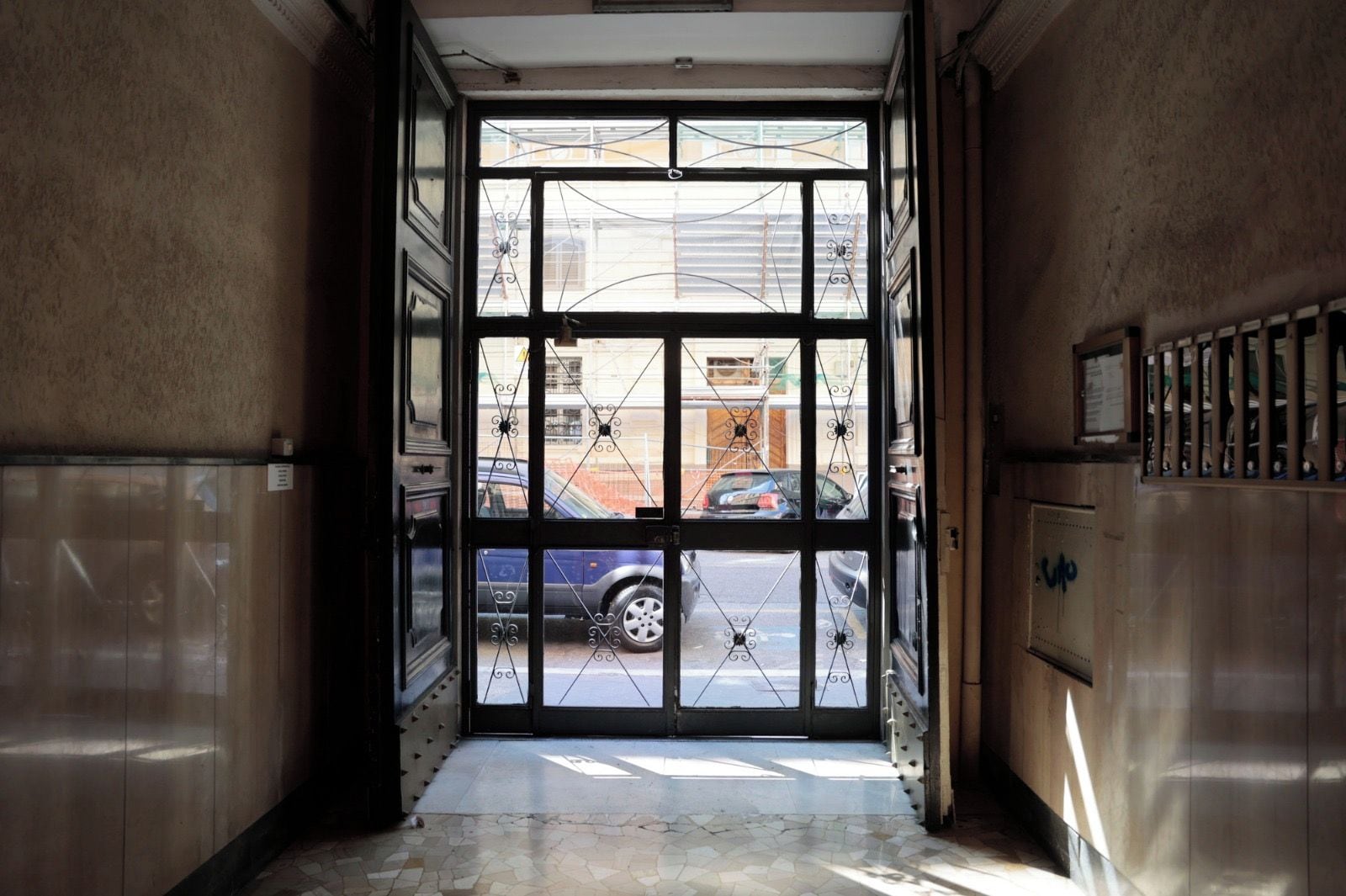 Un couloir intérieur avec une porte vitrée donnant sur un espace extérieur, un sol carrelé et une rambarde métallique sur le côté droit.