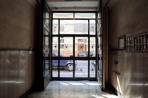 Un pasillo interior con una puerta de cristal que da a un área exterior, suelo de baldosas y barandilla metálica a la derecha.