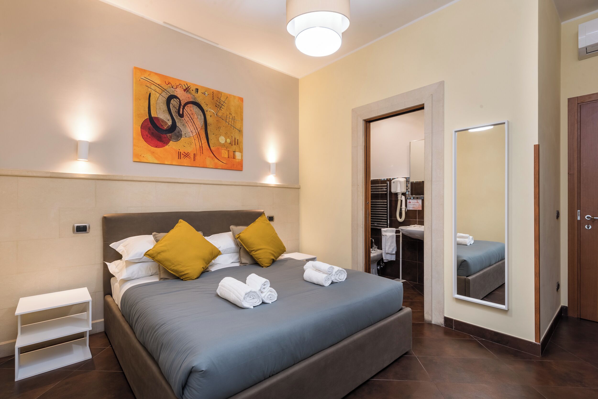 Ein ordentlich gemachtes Bett mit grauer Bettdecke und gelben Kissen, ein Nachttisch mit Lampe, ein Bild an der Wand und ein Spiegel, der den Raum reflektiert.