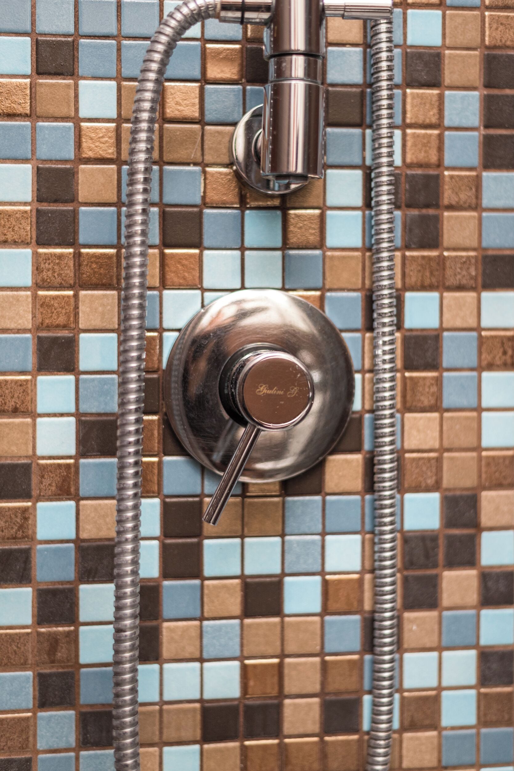 A shower with a chrome faucet and hose, set against a mosaic tile wall.