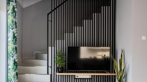 Un escalier moderne avec un téléviseur fixé sur un panneau noir orné de lignes verticales, une plante en pot sur la droite et un rideau vert sur la gauche.