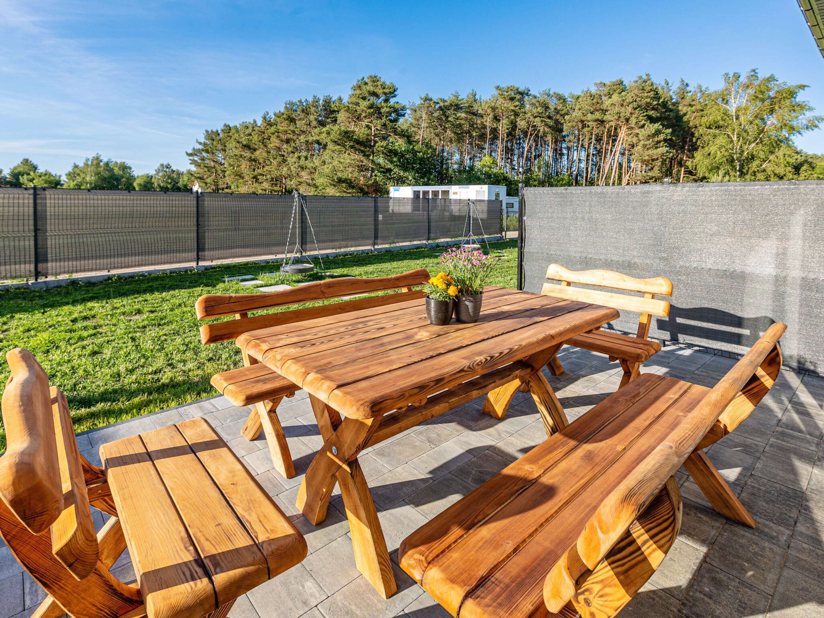 Un patio al aire libre con una mesa y bancos de madera, rodeado por una cerca y  árboles.