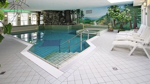Una piscina interior con agua cristalina, rodeada de sillas reclinables blancas y un mural que representa un paisaje forestal.