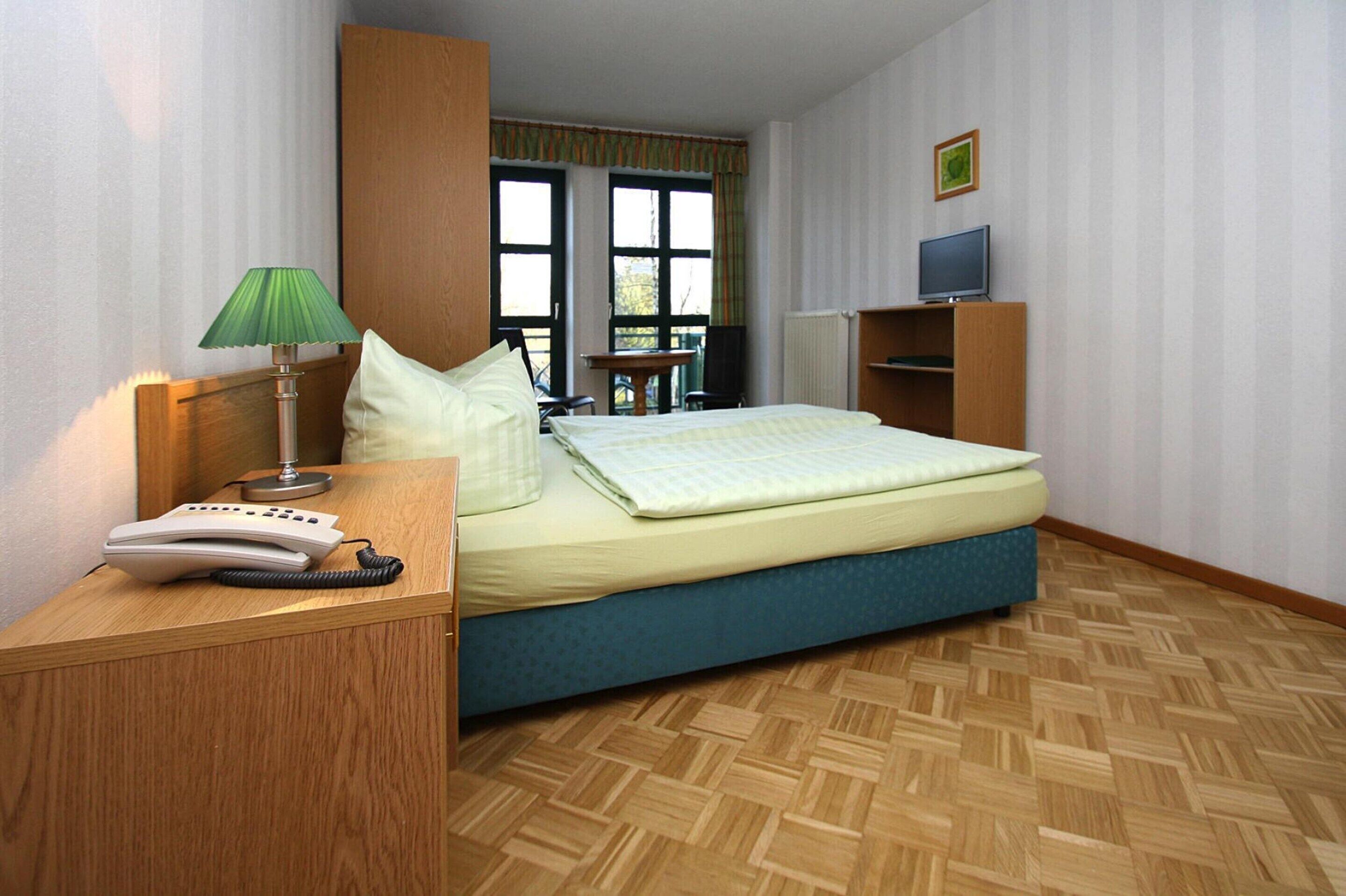 Ein Hotelzimmer mit Bett, Schreibtisch mit Telefon, Lampe, Fernseher auf einem Regal und einem Fenster mit Vorhängen.