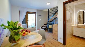 Un intérieur moderne avec un coin repas comprenant une table en bois, des chaises et un vase orné de fleurs et de fruits. On aperçoit un escalier au fond, menant à un niveau supérieur.