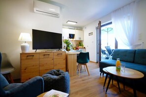 Een moderne woonkamer met een flatscreen televisie, een bank, een eettafel met fles en glas, en een keuken met kasten en een televisie.