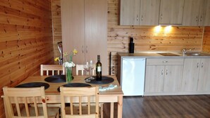 A wooden interior with a dining table, chairs, a vase with flowers, wine glasses, a bottle of wine, and a kitchen area with cabinets and a sink. - Holiday Houses for 4 People in Rewal-formerly TUI Ferienhaus (Rewal)