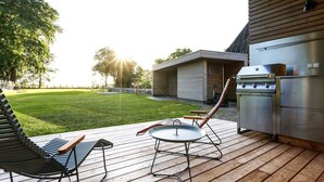 A deck with a grill and outdoor furniture, a grassy yard, and a building with a modern design. - Exklusives Landhaus auf Fehmarn (Fehmarn)