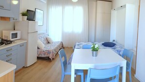 A compact kitchen with a microwave, stove, and cabinets. There's a dining area with a table, chairs, and a small potted plant. - Apartment in Bibione With Balcony (San Michele al Tagliamento)