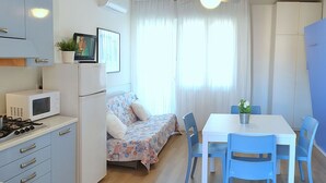 A compact kitchen with a microwave, stove, and refrigerator. There is a dining table with blue chairs and a bed with a floral bedspread. - Apartment in Bibione With Balcony (San Michele al Tagliamento)