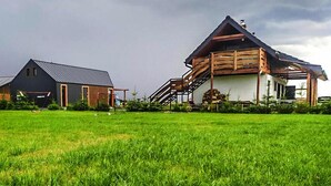 A rural setting with two wooden houses, one with a sloped roof and a balcony, and a large grassy field in the foreground. - Apartment in Jezierzany Near Lake Wicko (Postomino)