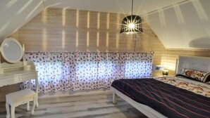 A bedroom with a sloped ceiling, a bed with a floral bedspread, a bedside table with a lamp, a small stool, and a window with patterned curtains. - Apartment in Jezierzany Near Lake Wicko (Postomino)