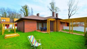 A backyard with a brick house, a wooden swing set, a colorful chair, and a pergola. - Spacious Holiday Home With Private Garden (Mielno)