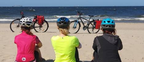 Three individuals seated on a sandy beach with bicycles and helmets, overlooking the ocean.