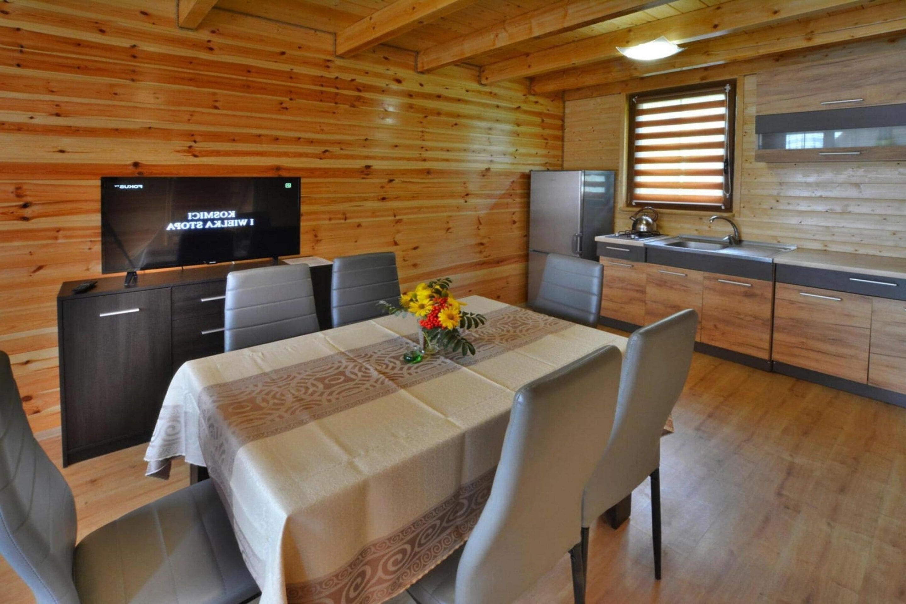 A wooden interior with a dining table set for four, a kitchen area with a refrigerator and sink, and a flat-screen TV mounted on the wall.
