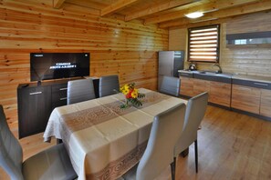 A wooden interior with a dining table set for four, a kitchen area with a refrigerator and sink, and a flat-screen TV mounted on the wall. - Comfortable Holiday Home in a Peaceful Setting (Postomino)