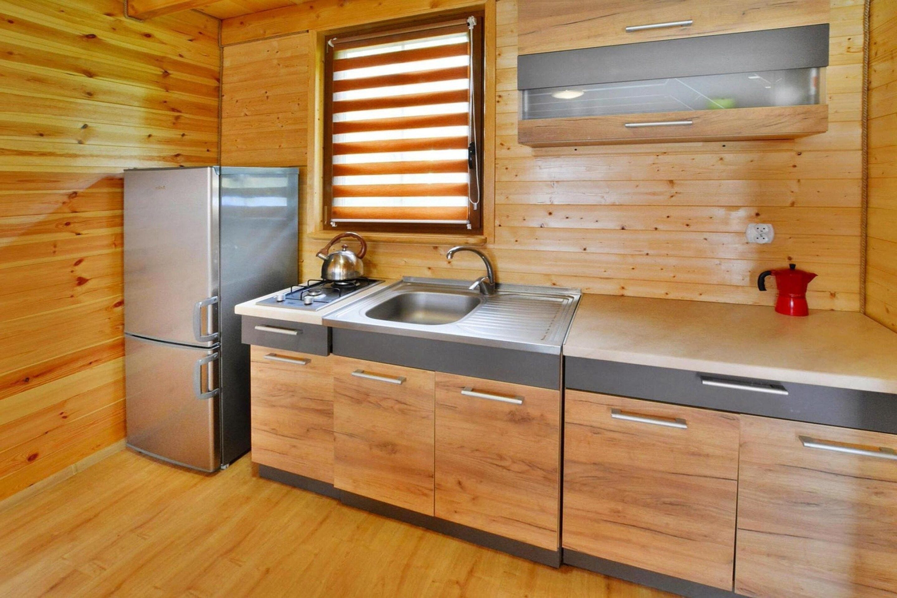 A compact kitchen with wooden walls, a stainless steel refrigerator, a sink, and a kettle.