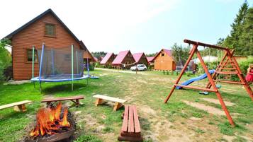 A wooden cabin with a trampoline, a swing set, and a fire pit in the yard.