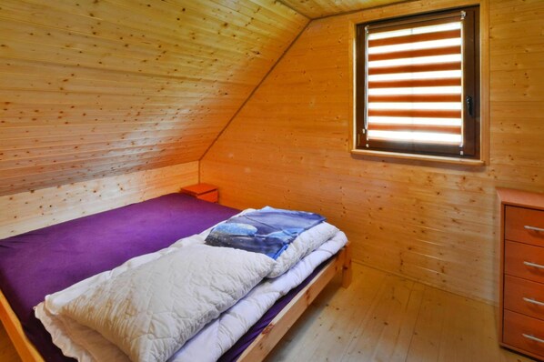A small, cozy room with a sloped ceiling, a single bed with a purple and white bedspread, a small wooden nightstand, and a window with blinds. - Holiday House Jaroslawiec Near Tropicana Park (Postomino)