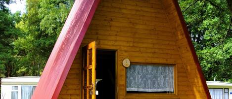 A triangular wooden cabin with a door open, a window with lace curtain, and a small porch area.