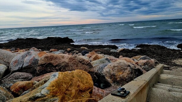 A rocky shoreline with waves crashing against the rocks under a cloudy sky.