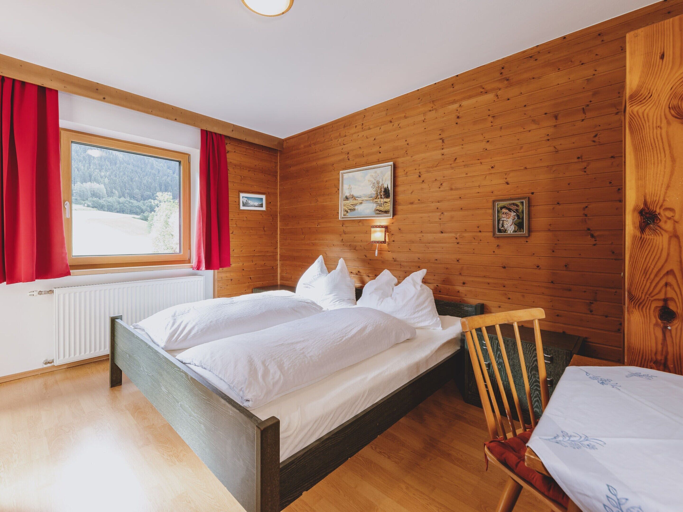 Une chambre aux murs lambrissés, avec un lit, une chaise et une fenêtre ornée de rideaux rouges.
