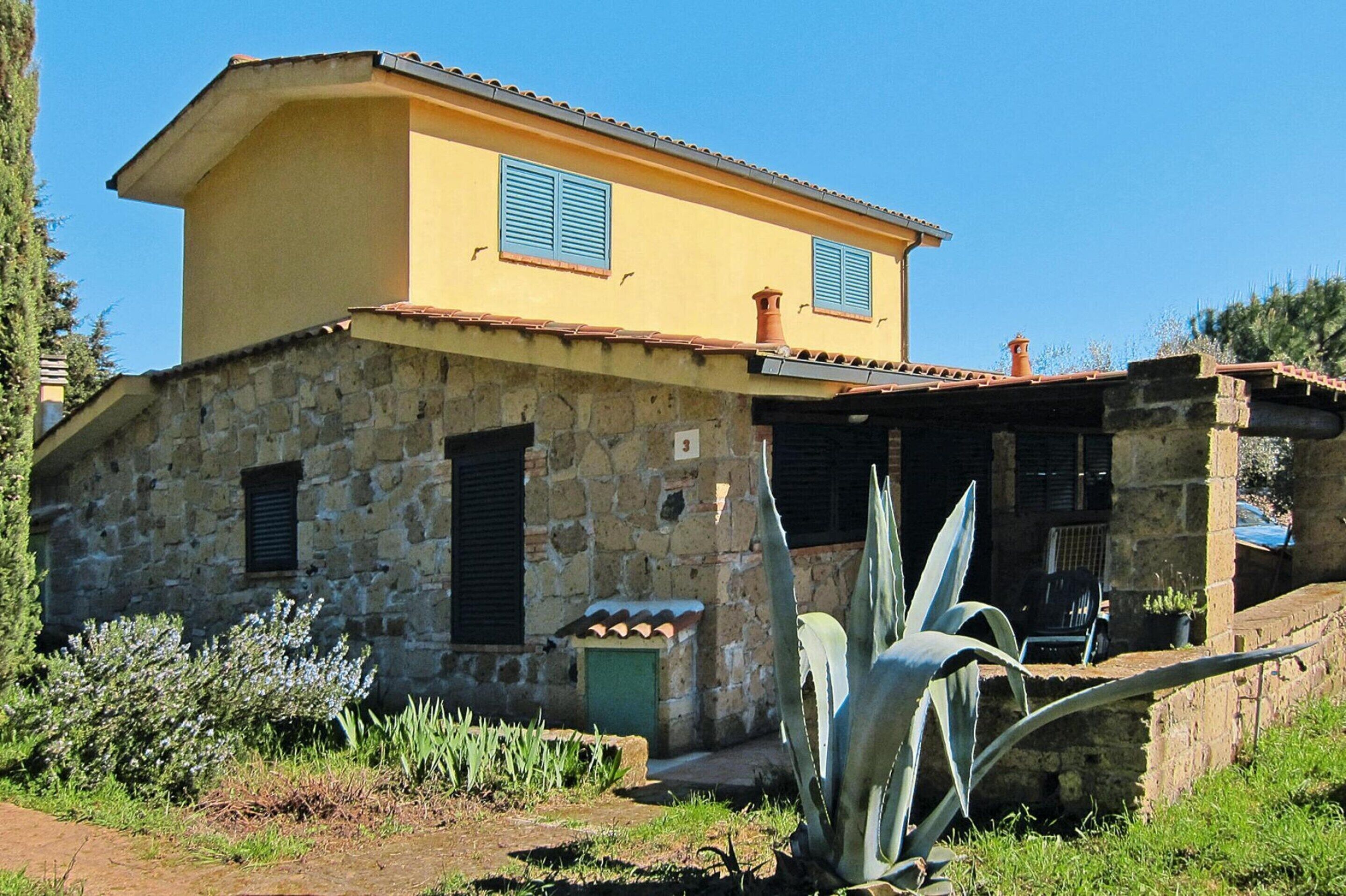 Una casa de piedra de dos plantas con fachada amarilla y contraventanas azules, que incluye una entrada con muro de piedra y un pequeño patio.