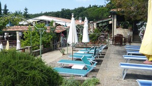 Un espace aménagé au bord de la piscine, avec des chaises longues et des parasols, entouré de verdure et bordé d’un bâtiment dont le toit est recouvert de tuiles.