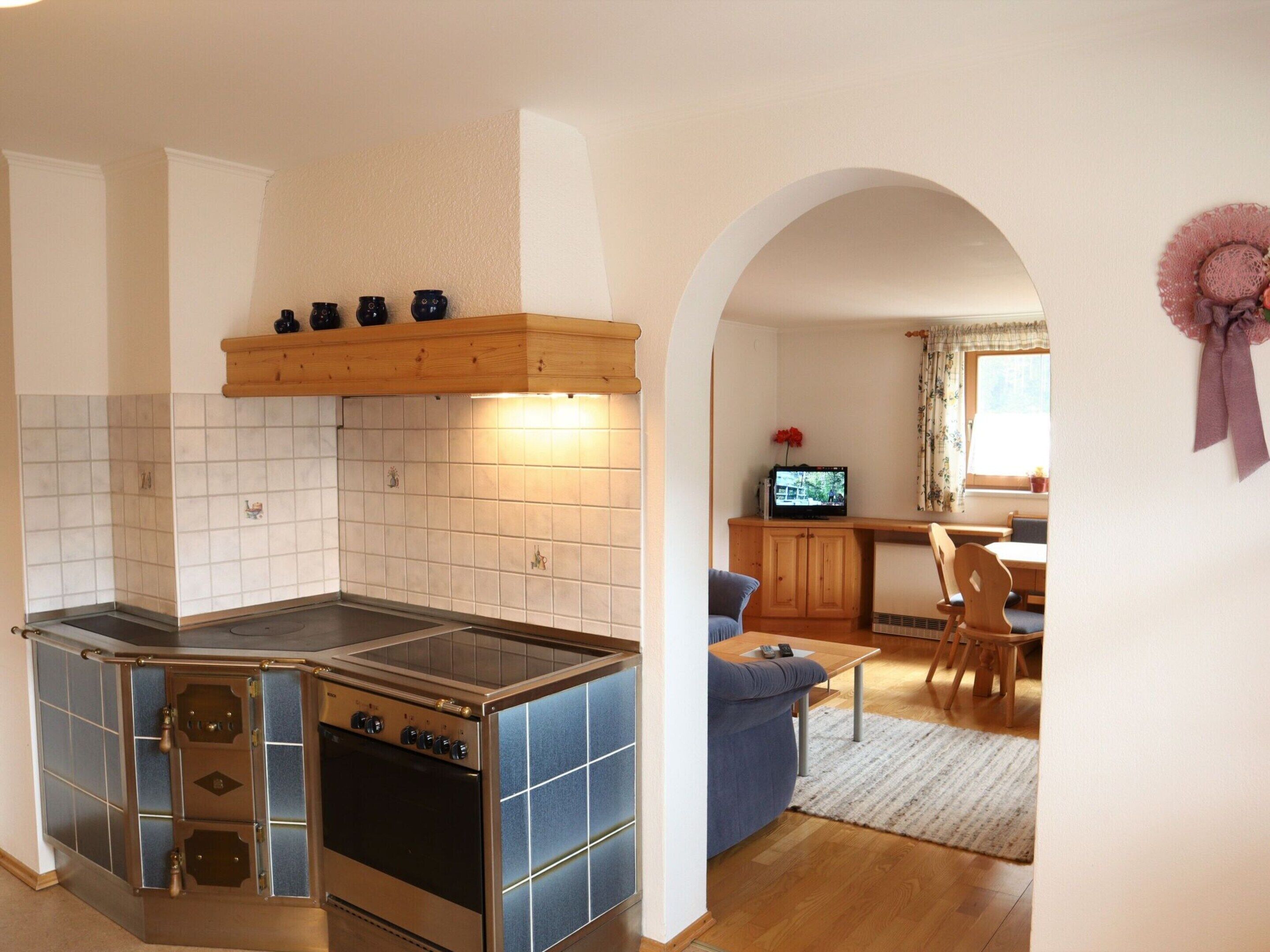 Una cucina compatta con piano cottura, forno e una mensola in legno. In secondo piano, si vede un'area pranzo con una sedia blu e un tavolo.