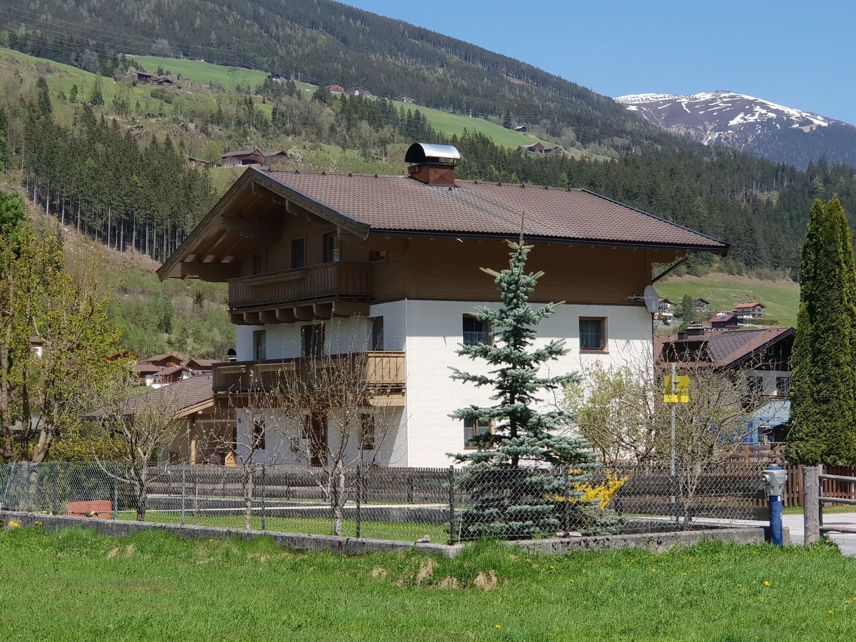 A chalet-style house with a sloped roof, wooden balconies, and a fenced yard with a coniferous tree.