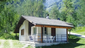 A small, single-story house with a tiled roof, a porch with chairs, and a view of a forested mountain area. - Apartment Santa Lucia in Bezecca (Ledro)