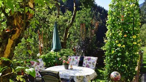 A round table with a floral tablecloth, surrounded by wicker chairs, in a garden with a wooden trellis and potted plants. - Apartment in Huttau Near Ski Amade Slopes (Huettau)