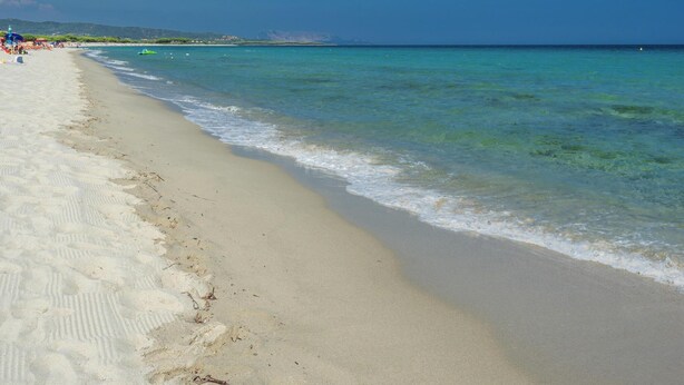 Une plage à l’eau turquoise et limpide, bordée d’un rivage sablonneux, avec des collines qui se dessinent à l’horizon.