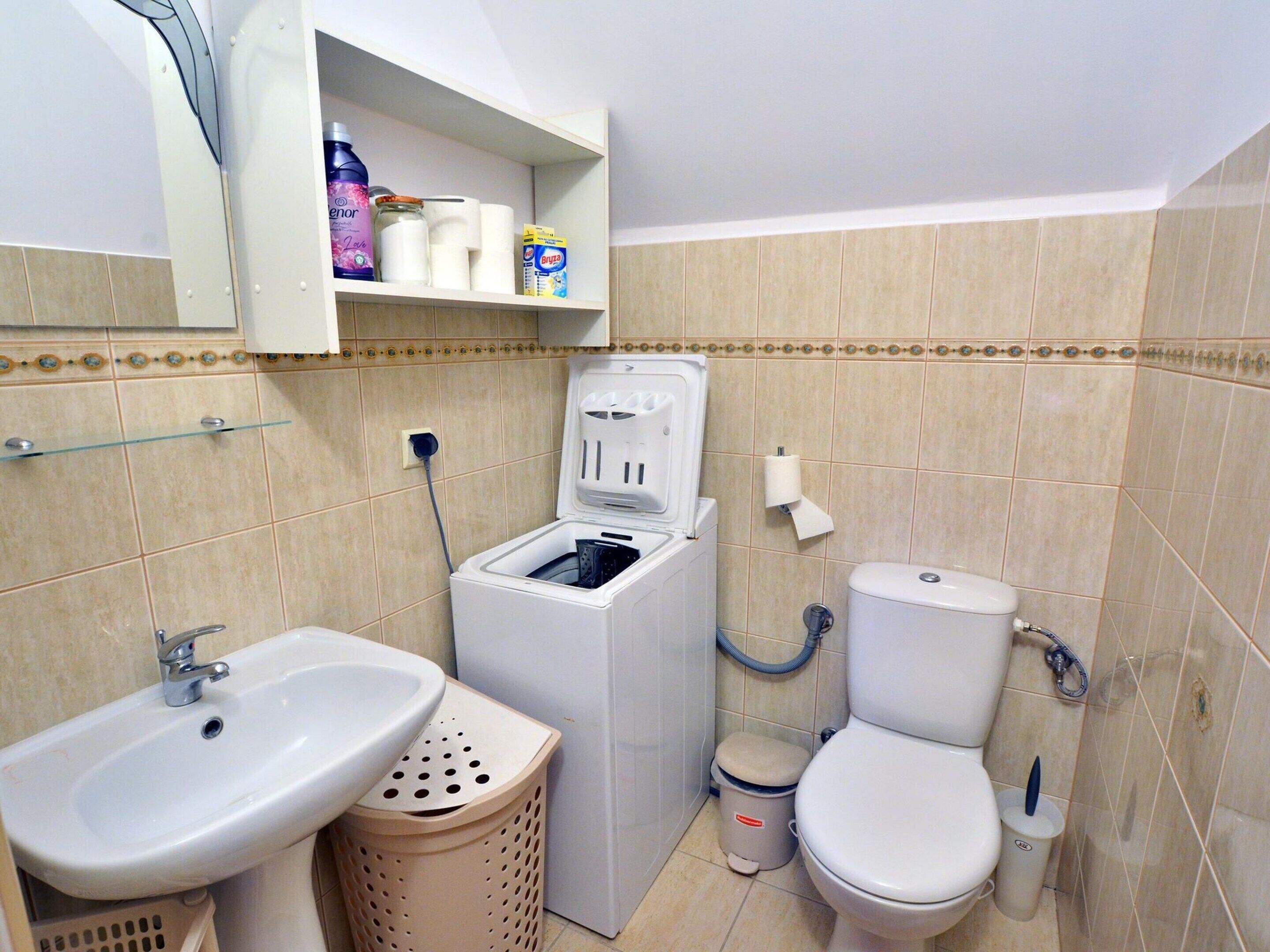 Une petite salle de bain comprenant un lavabo, un lave-linge, des toilettes et une étagère avec des articles de toilette.