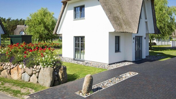 A white house with a thatched roof, a stone wall, and a flower bed with red flowers. - Komfortables Ferienhaus mit Sauna in Fuhlendorf (Fuhlendorf)