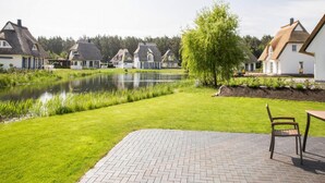 A serene residential area with thatched-roof houses, a pond, and a paved patio with a chair. - Komfortables Ferienhaus mit Sauna in Fuhlendorf (Fuhlendorf)