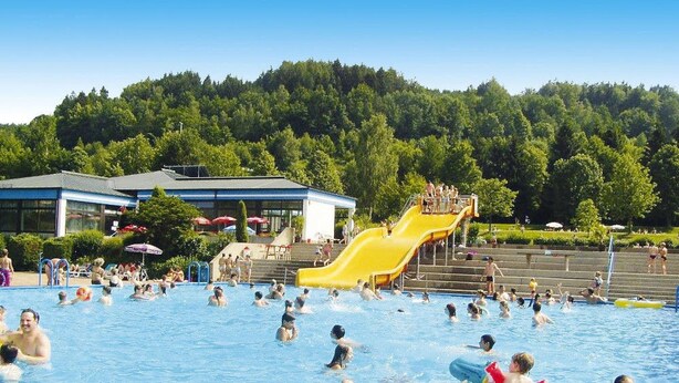 Ein öffentliches Schwimmbecken mit einer gelben Rutsche und einem bewaldeten Hügel im Hintergrund.