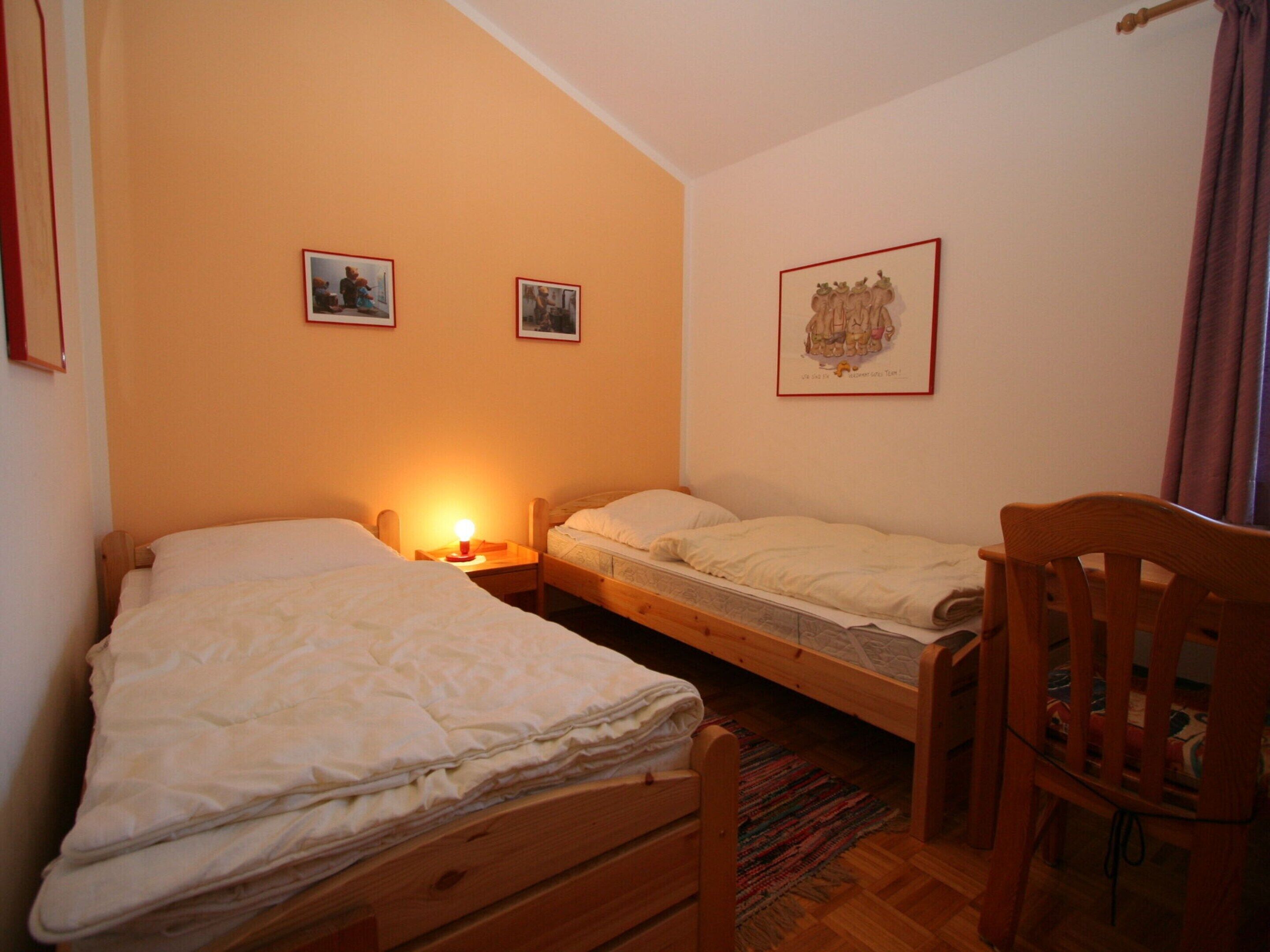 A small, cozy bedroom with two single beds, a wooden chair, and a nightstand with a lamp.
