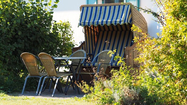 Un patio con sedie in vimini e un tenda a righe, immerso nel verde e affiancato da un edificio con persiane blu.