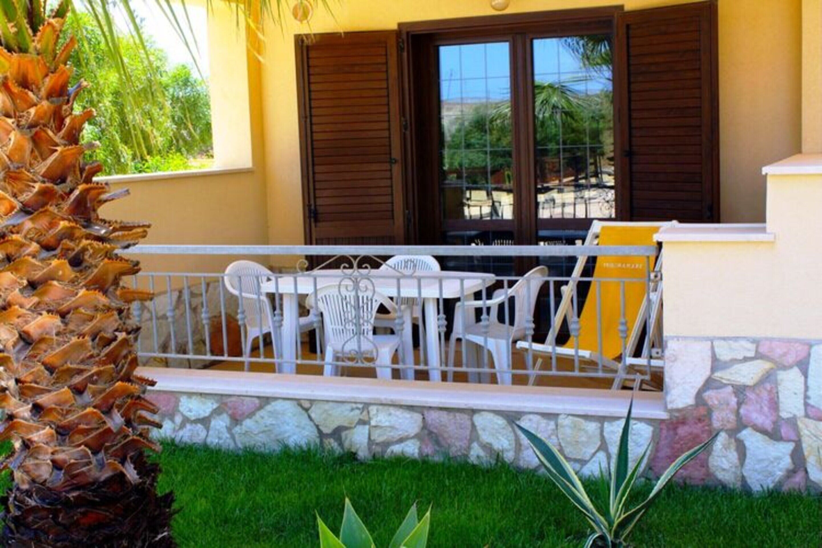 A patio with white chairs and a table, a stone wall, and a palm tree.
