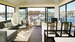 A modern living room with a sofa, coffee table, and dining area with chairs, overlooking a marina with boats.